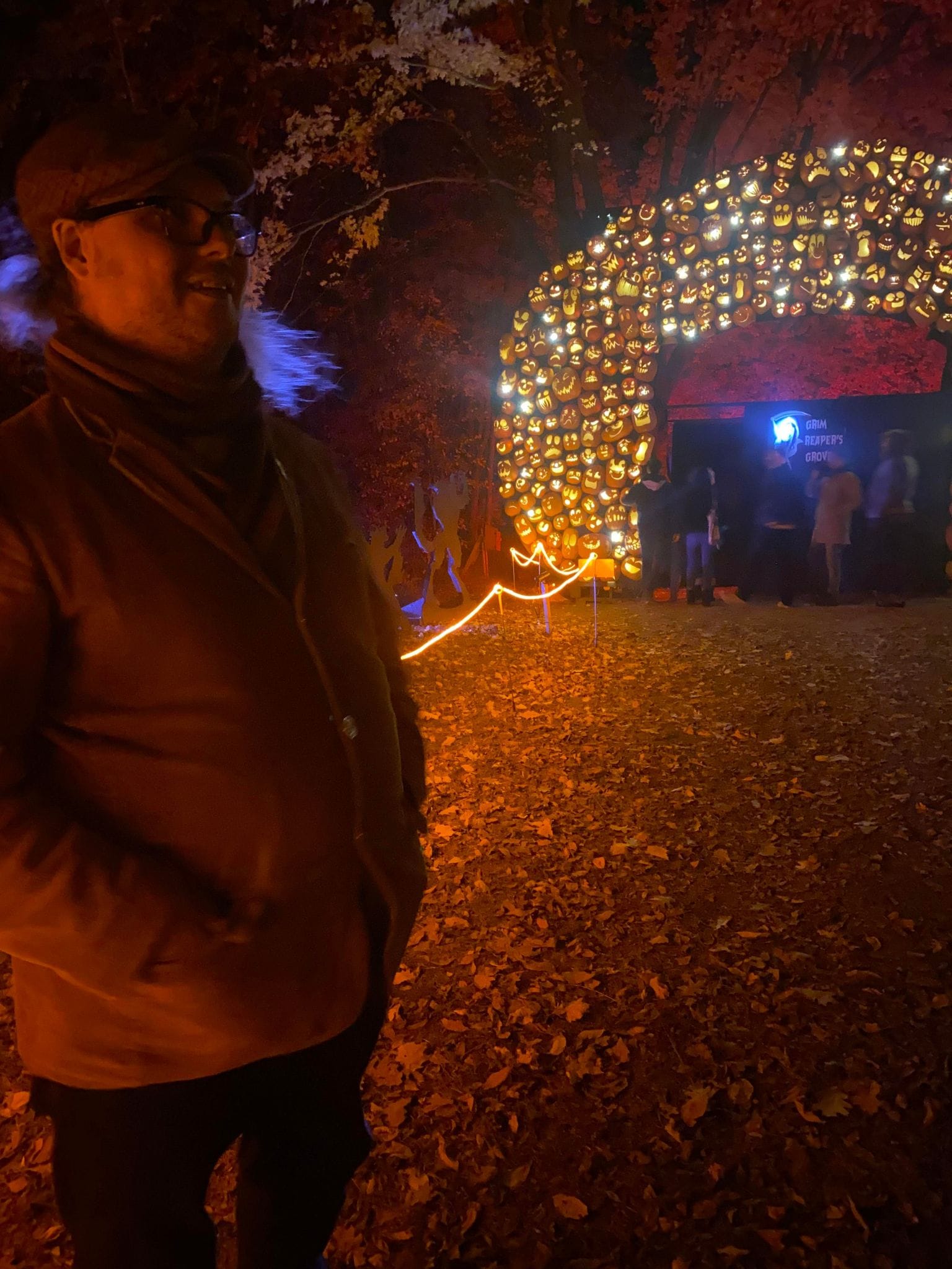 A man stands in front of an arch made of jack-o-lanturns, his hair looks purple in the light, Photo 1